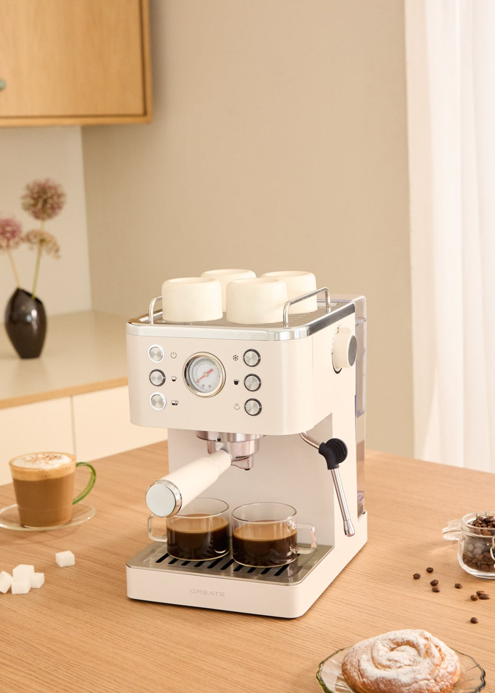 Una máquina de espresso de color crema en una mesa de madera con dos tazas de café, pasteles y granos de café.