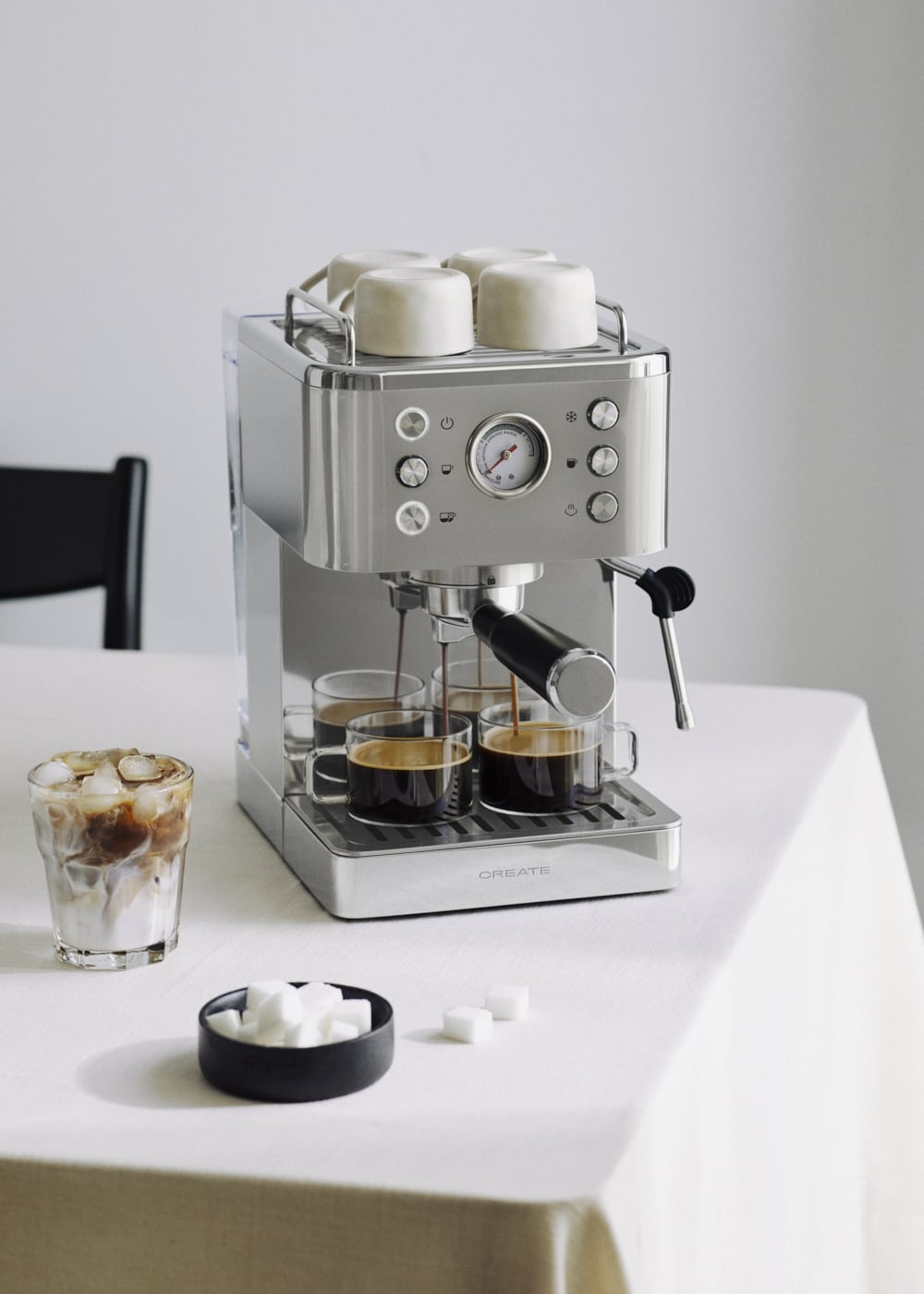 Una máquina de café plateada prepara café en cuatro vasos, con café helado y terrones de azúcar al lado.