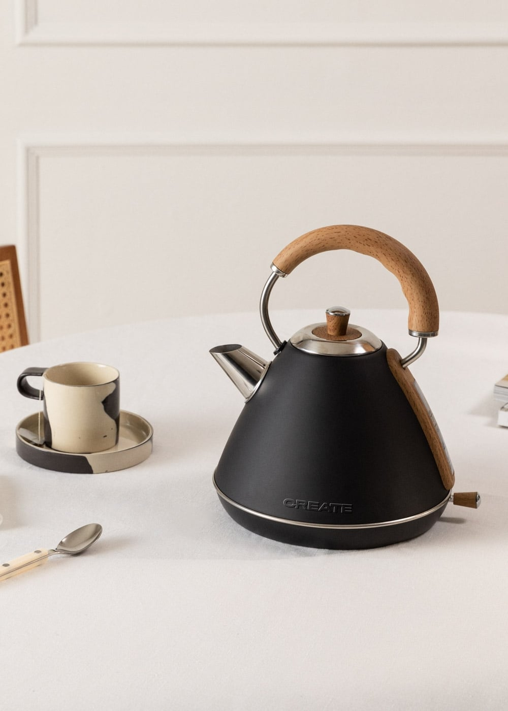Un hervidor negro con asa de madera descansa sobre una mesa junto a una taza y una cuchara.