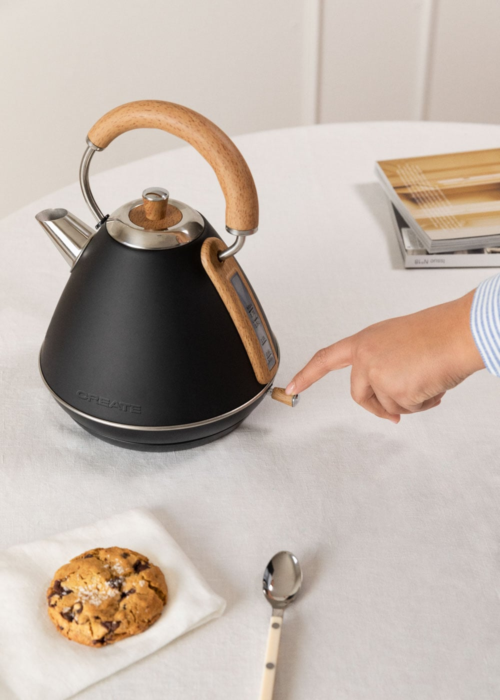 Una mano enciende un hervidor negro con asa de madera. Una galleta y una cuchara están sobre la mesa.