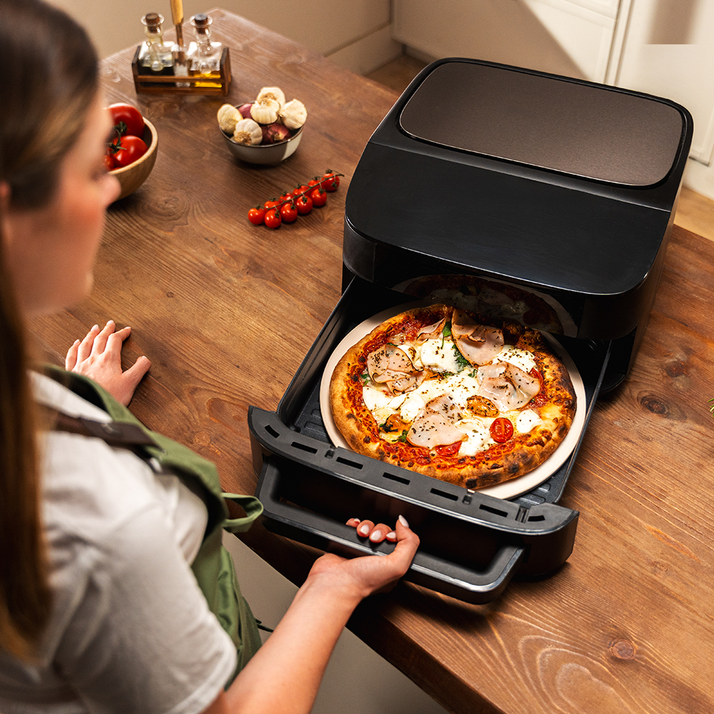 Una donna apre il cassetto di una friggitrice ad aria nera con dentro una pizza appena cotta.