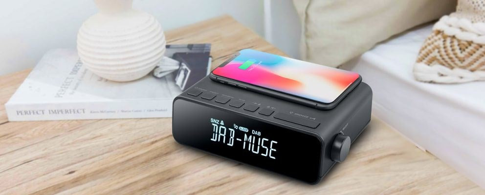 A black digital clock radio on a wooden table with a smartphone charging on top.
