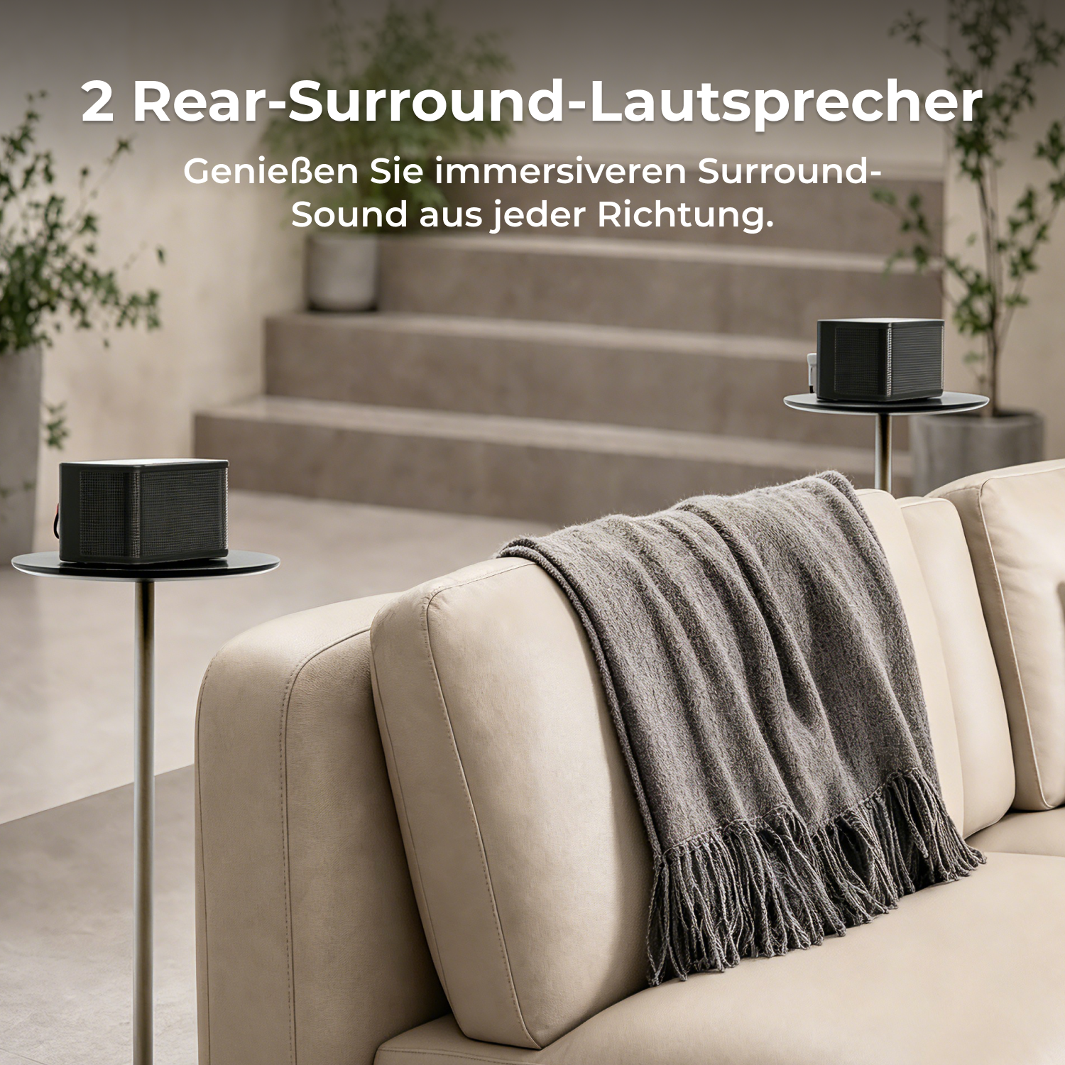 Zwei graue Surround-Lautsprecher stehen auf Ständern neben einem beige Sofa mit einer Decke.