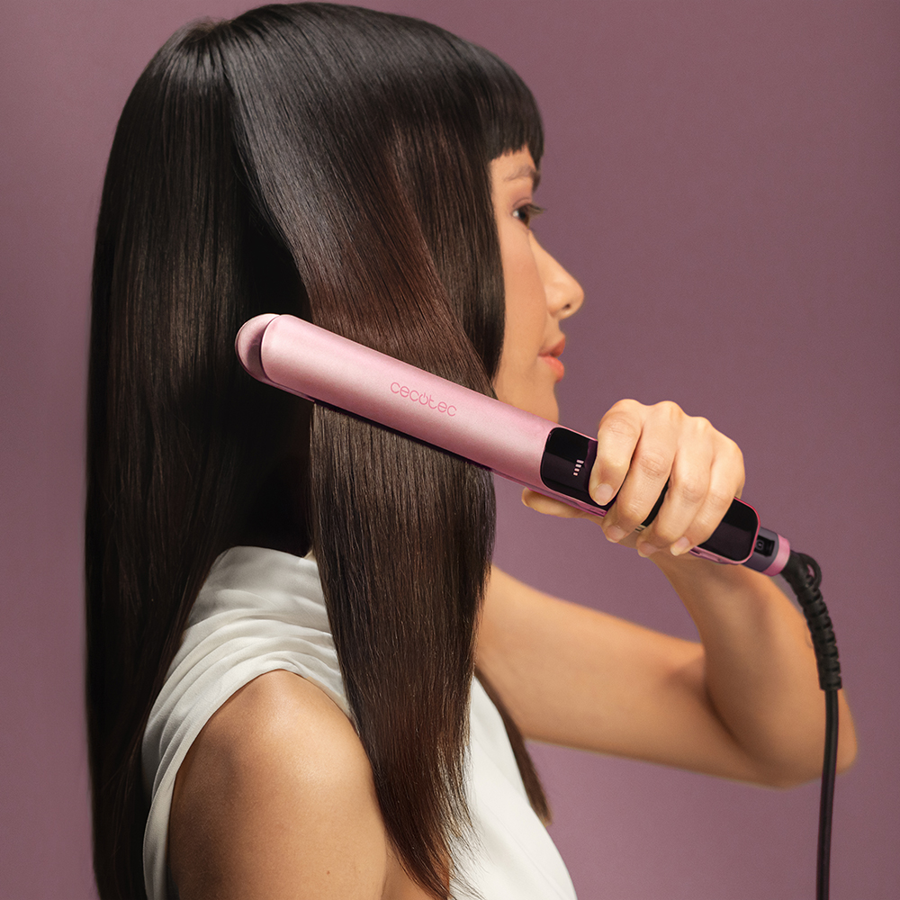 Una donna con i capelli scuri liscia una ciocca di capelli con una piastra rosa su sfondo viola.