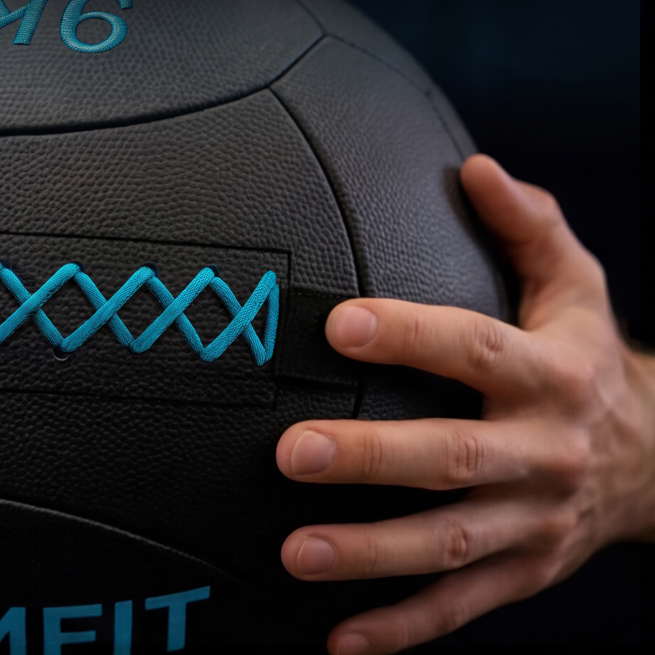 Una mano sostiene un balón medicinal negro con costuras azules y letras azules sobre fondo oscuro.