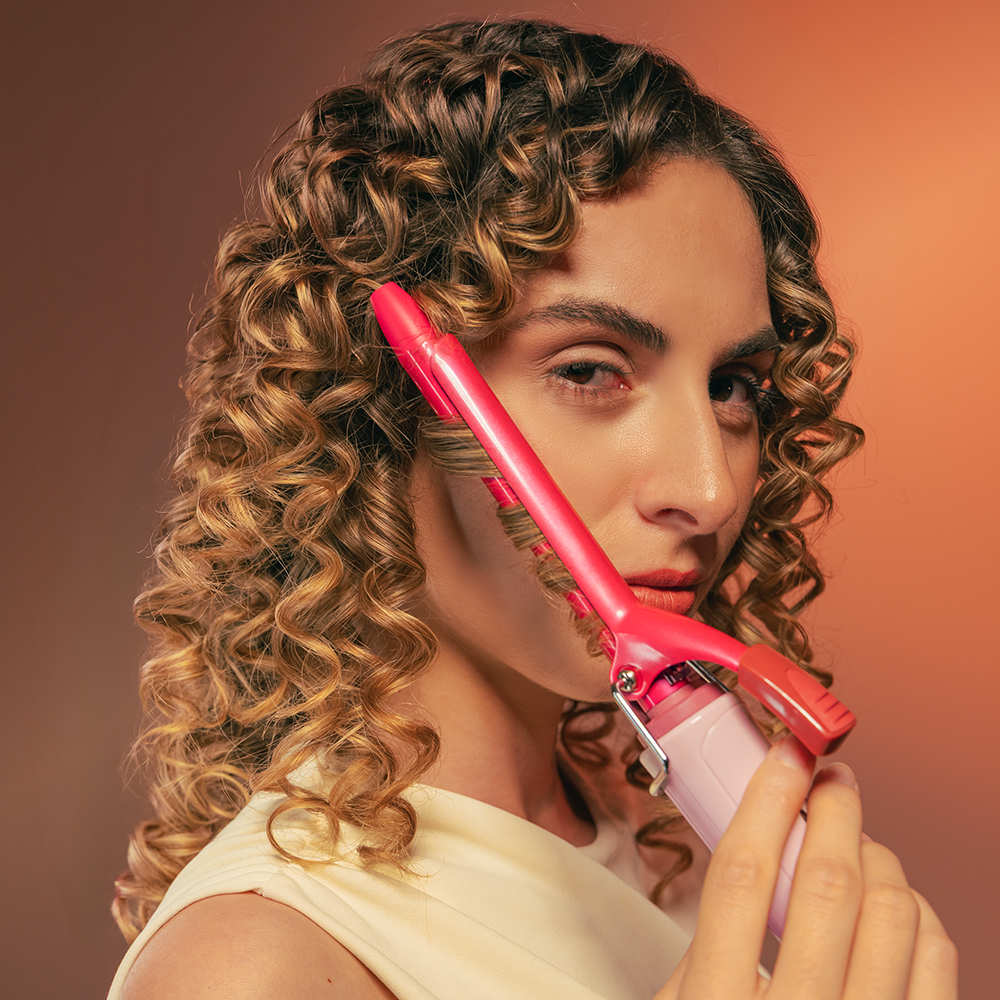 Una donna con capelli ricci arriccia una ciocca con un ferro rosa su sfondo arancione.