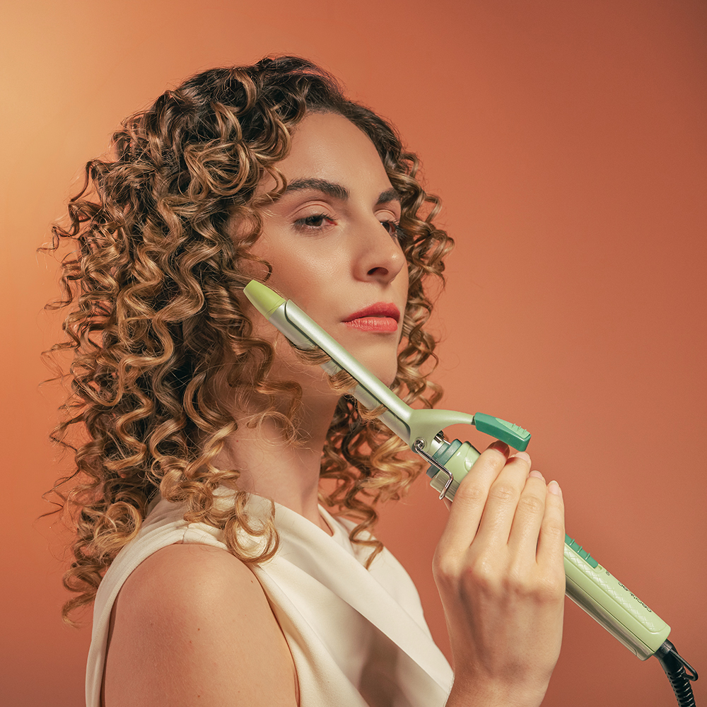 Una donna dai capelli castani ricci usa un ferro arricciacapelli verde chiaro su sfondo arancione.