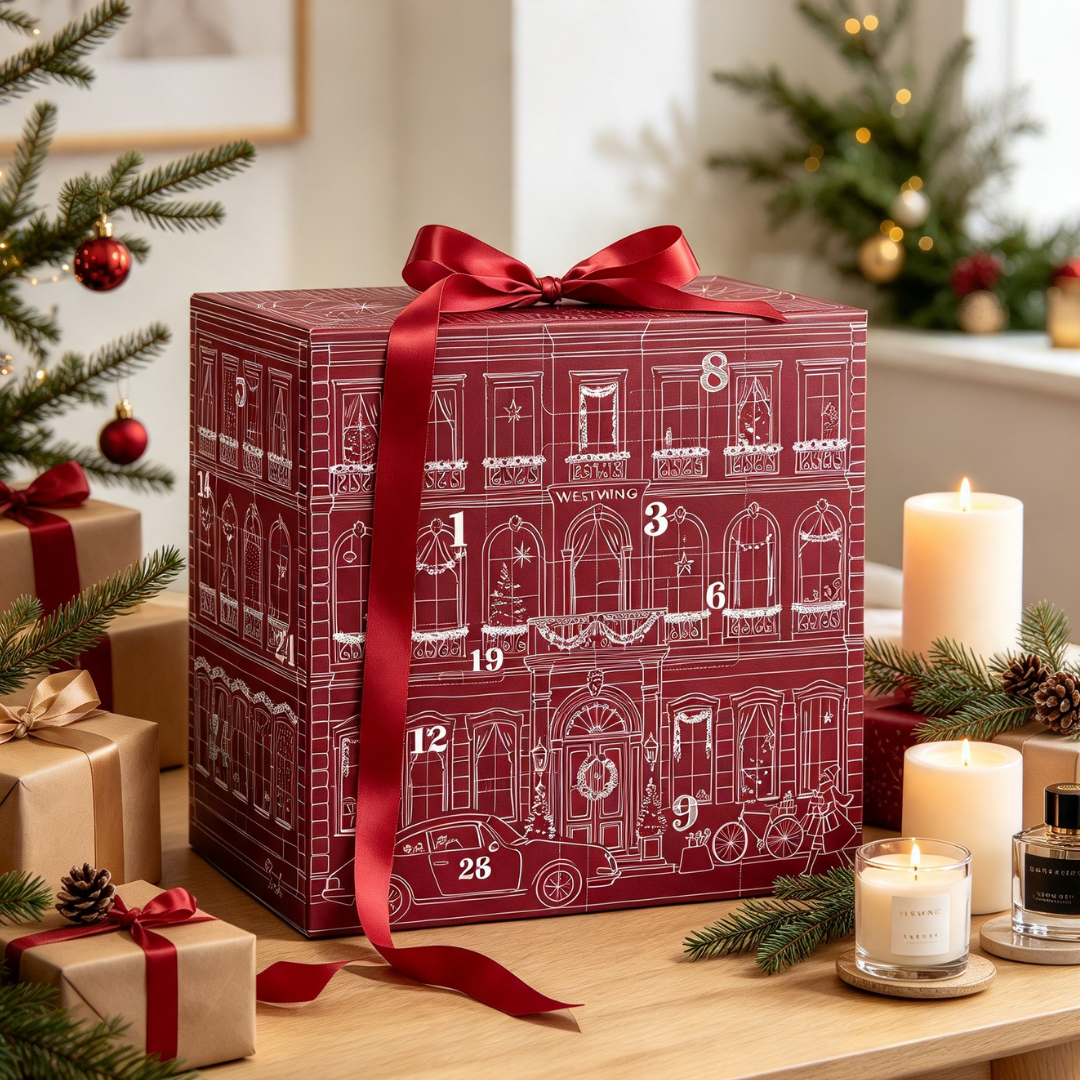 Eine große rote Adventskalenderbox mit Satin Schleife steht auf einem Holztisch, umgeben von Kerzen und Geschenken.