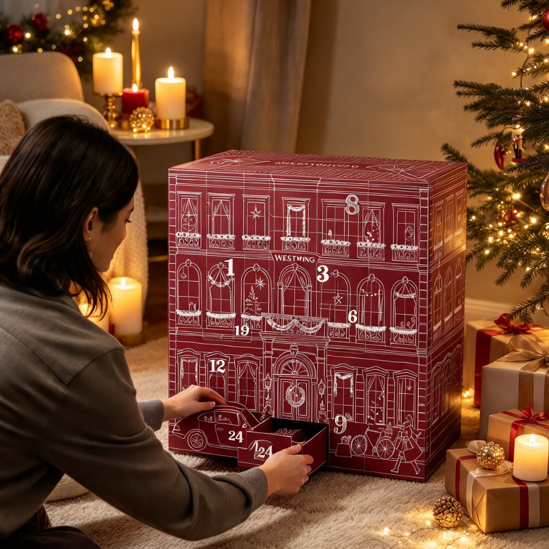 Eine Frau öffnet einen großen roten Adventskalender mit weißer Gebäudezeichnung vor einem Weihnachtsbaum.