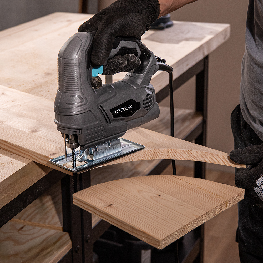 Una mano con guante usa una sierra caladora gris para cortar una tabla de madera en un banco de trabajo.