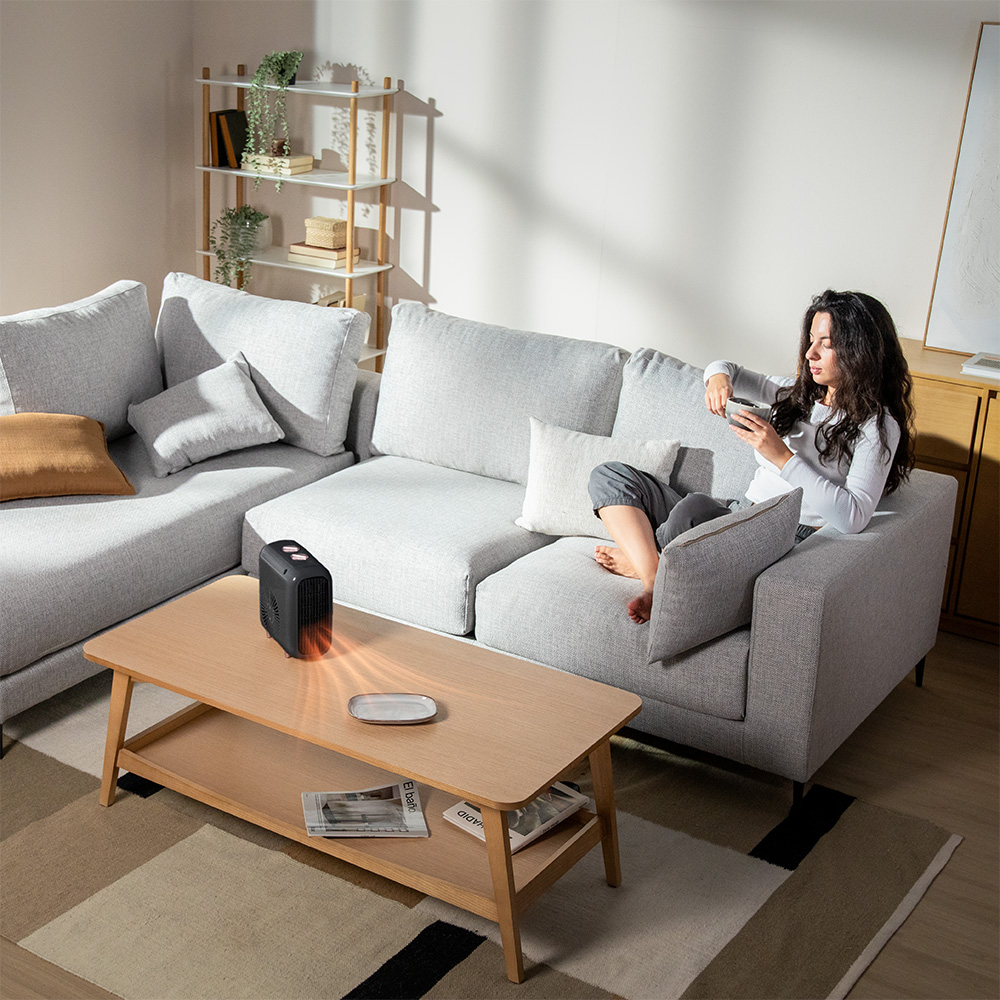 Eine Frau sitzt auf einem grauen Sofa, während auf einem Holztisch ein kleiner schwarzer Heizlüfter steht.