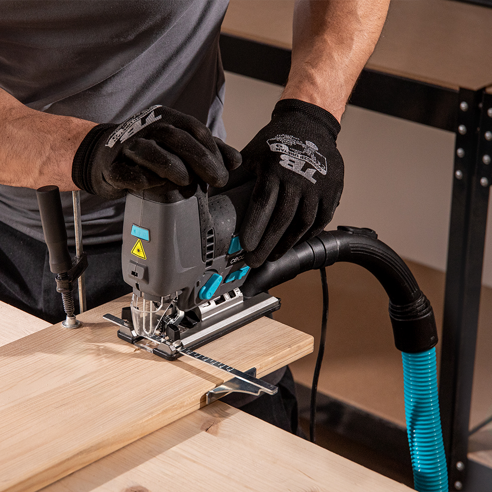 Una persona utiliza una sierra de calar gris con una manguera azul conectada para cortar una tabla de madera.