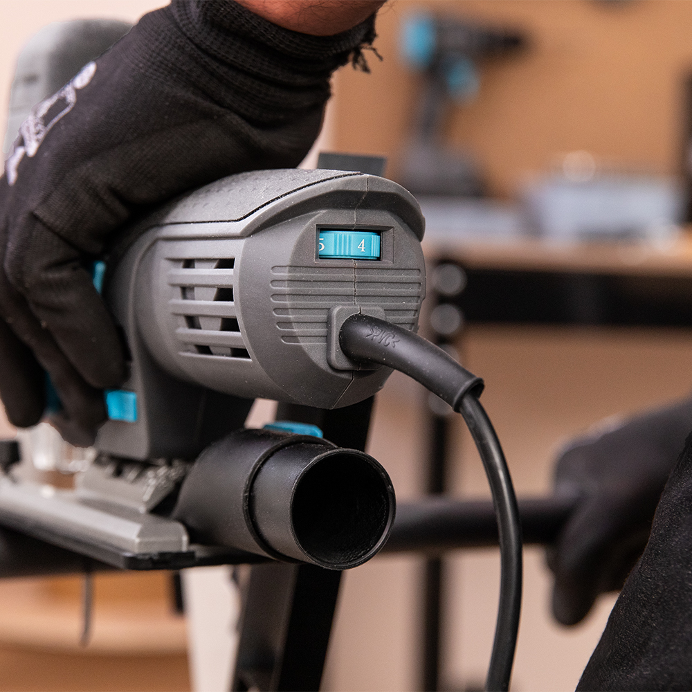 Una mano con guante sostiene una sierra caladora gris con cable de alimentación y control de velocidad.