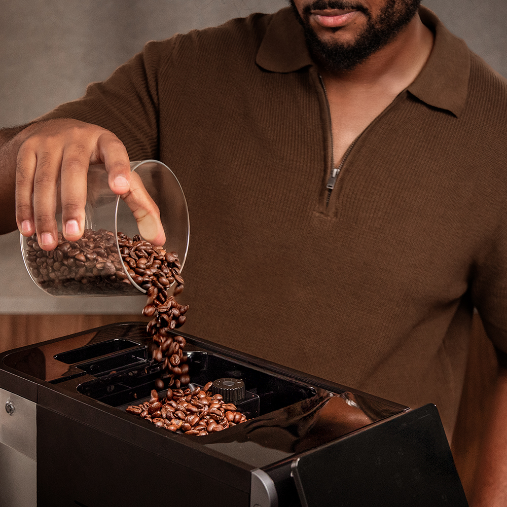 Ein Mann in einem braunen Polohemd gießt Kaffeebohnen aus einem Glas in eine Kaffeemaschine.
