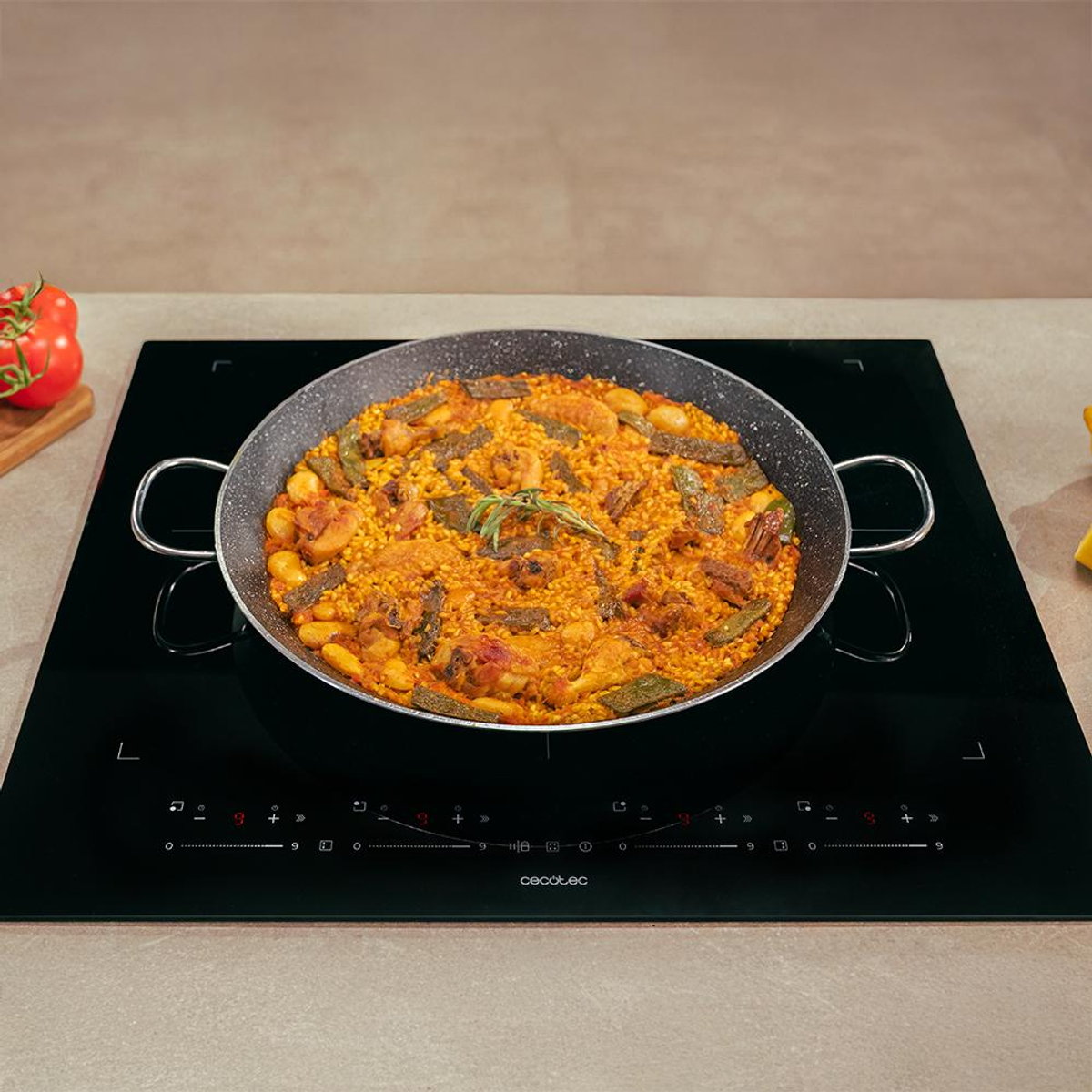 Una sartén de paella con arroz, carne y verduras está sobre una encimera de inducción negra con controles táctiles.