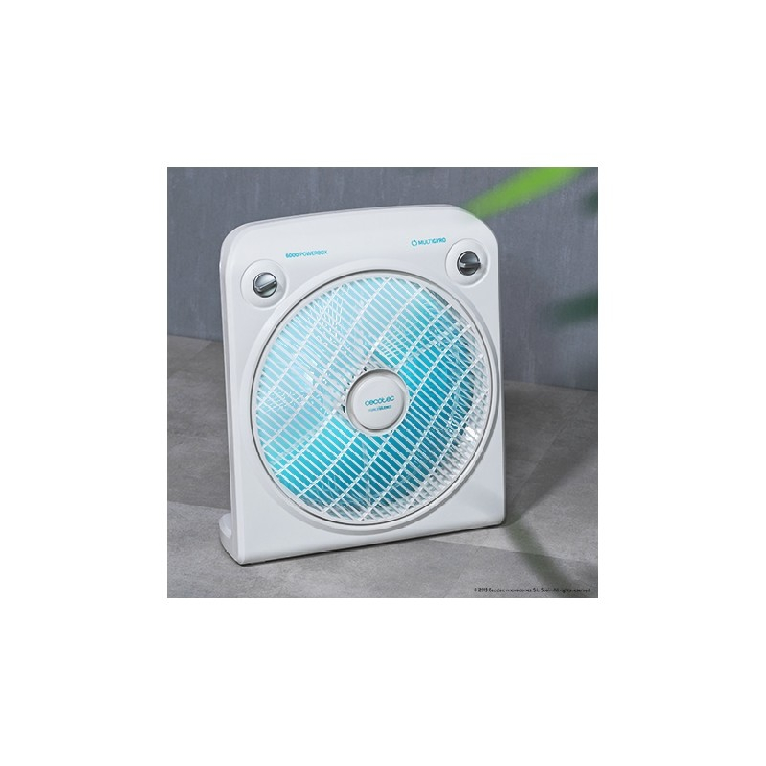 Un ventilador de caja blanco con rejilla interior azul sobre una superficie gris ante una pared.