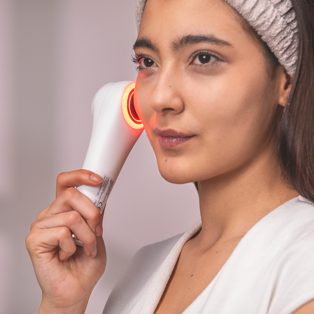 Una mujer usa un dispositivo facial blanco con luz roja en su mejilla.