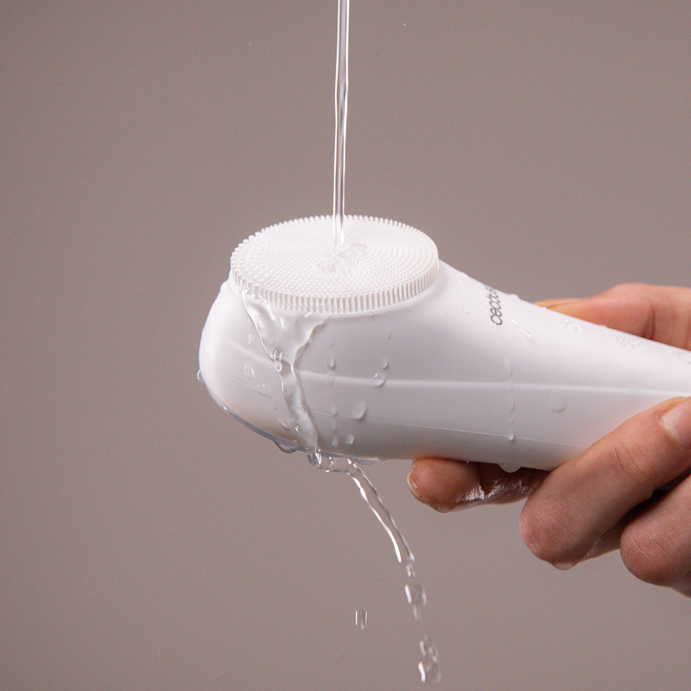 Una mano sostiene un cepillo limpiador facial blanco bajo el agua corriente con fondo liso.