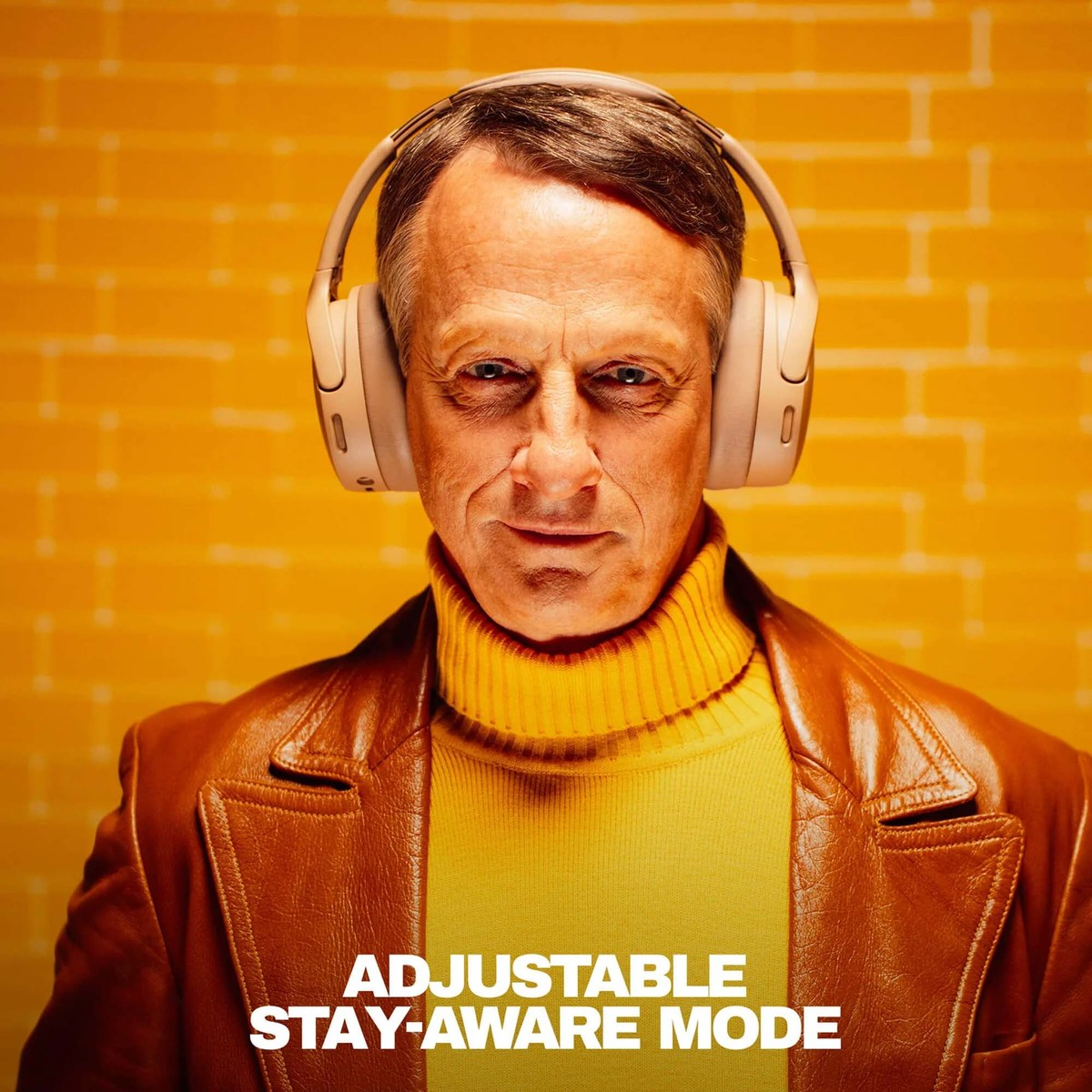 Un hombre mayor con auriculares, jersey de cuello alto amarillo y chaqueta marrón sobre fondo amarillo.