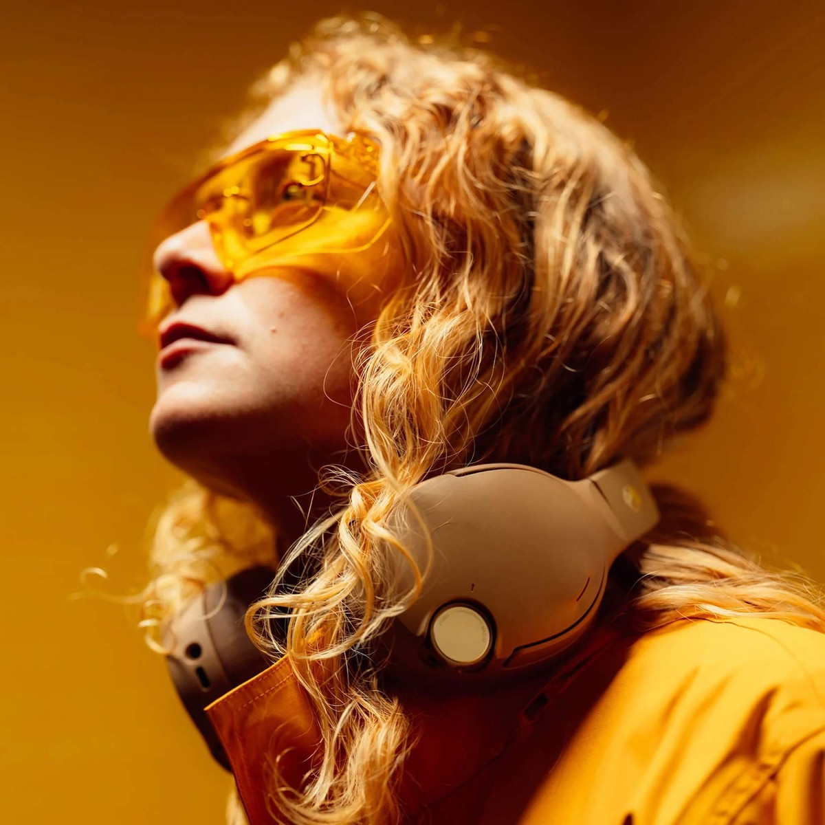 Una persona con cabello rubio rizado, gafas protectoras amarillas y auriculares al cuello sobre fondo amarillo.