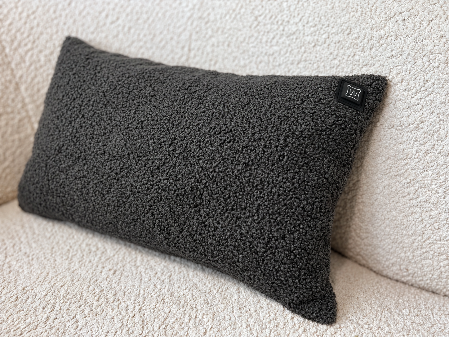 Ein dunkelgraues, flauschiges Kissen mit einem kleinen schwarzen Etikett liegt auf einer cremefarbenen Couch.