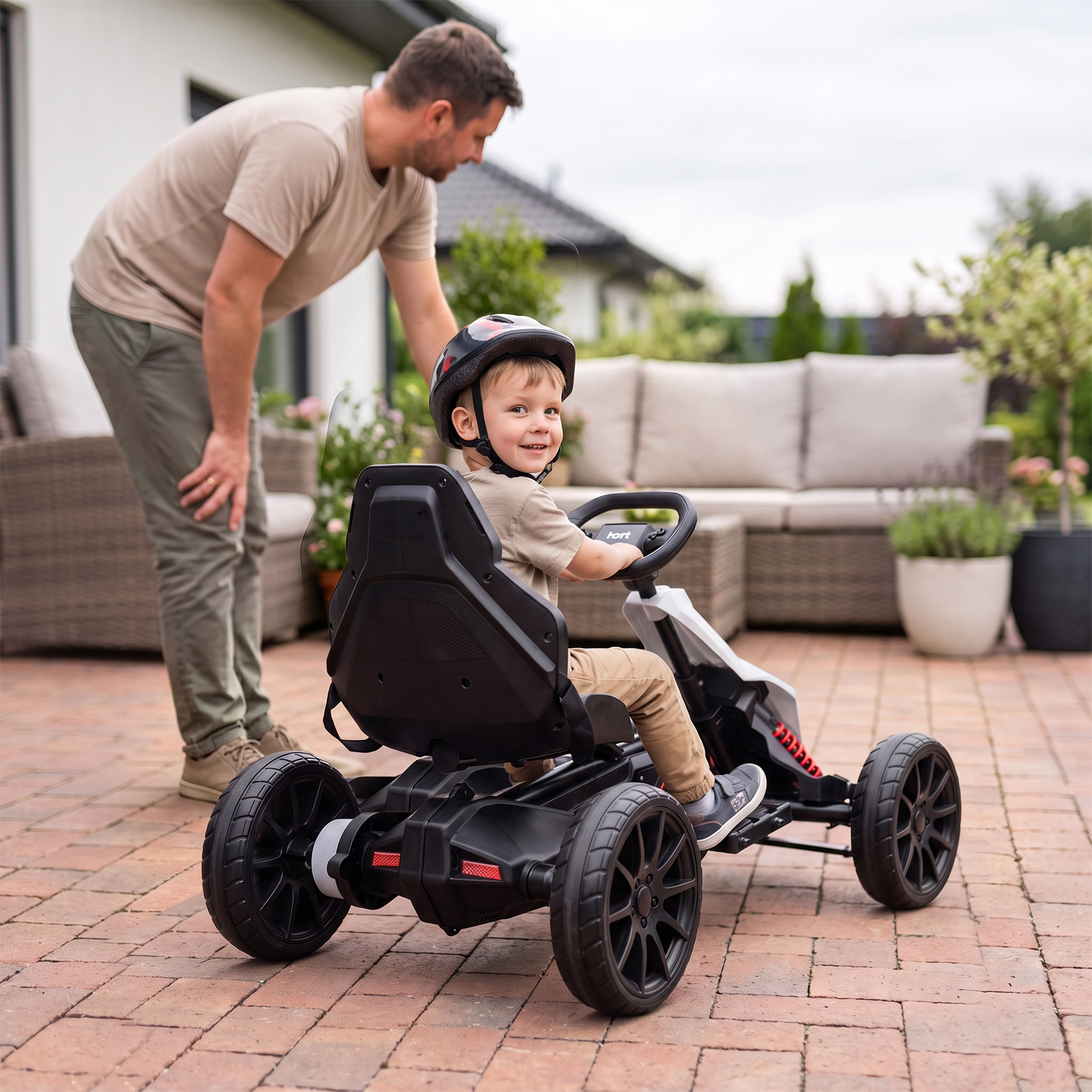 Un niño feliz con casco conduce un kart en un patio adoquinado mientras su padre está detrás de él.