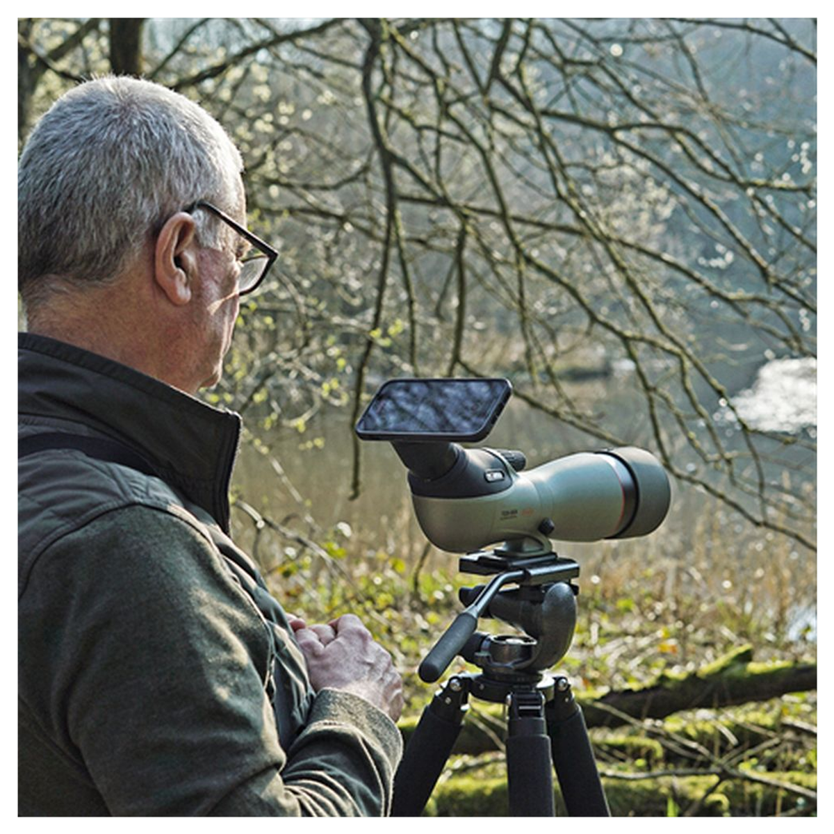 Ein älterer Mann mit Brille blickt im Wald durch ein auf einem Stativ montiertes Spektiv.