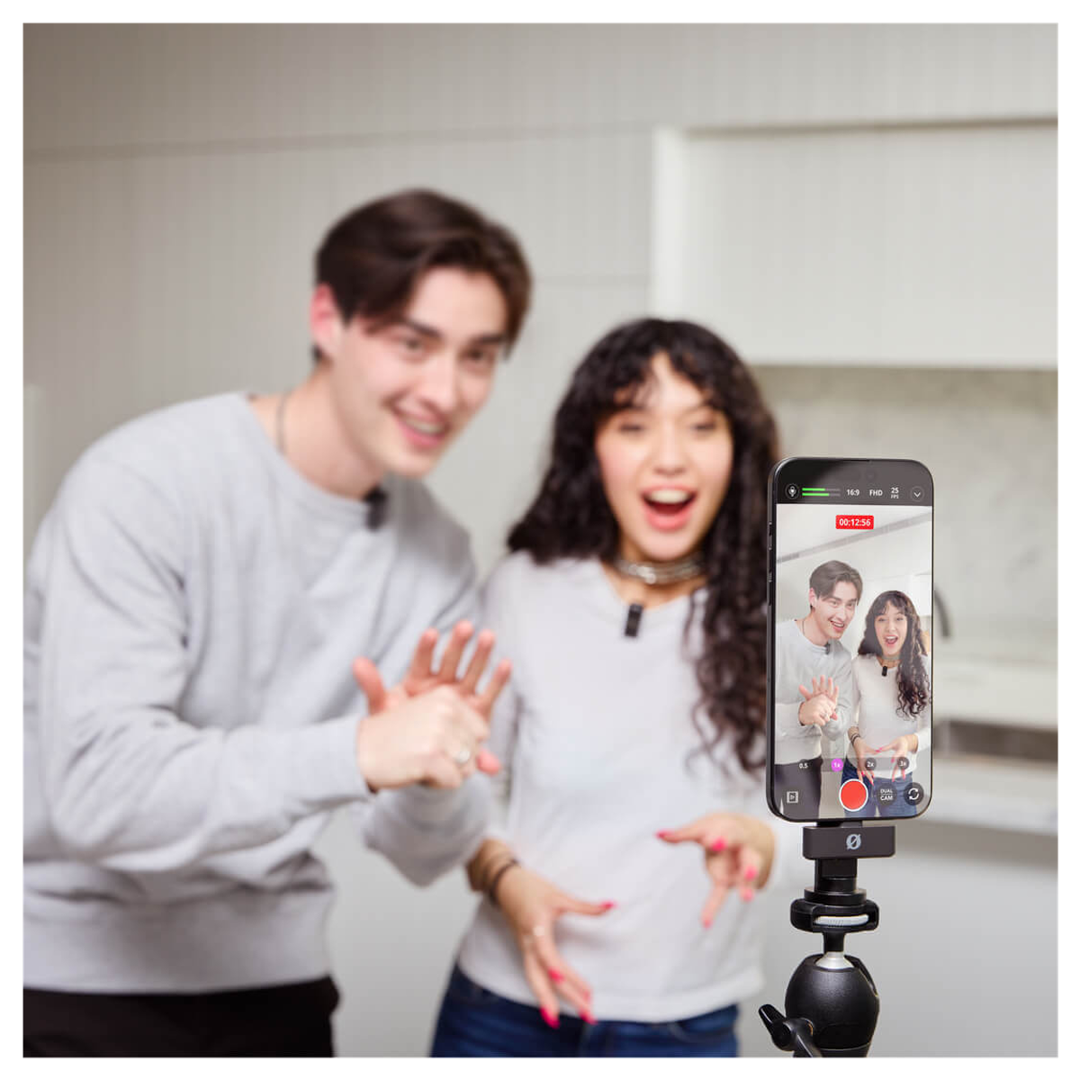 A young couple enthusiastically waves at a smartphone camera mounted on a tripod.