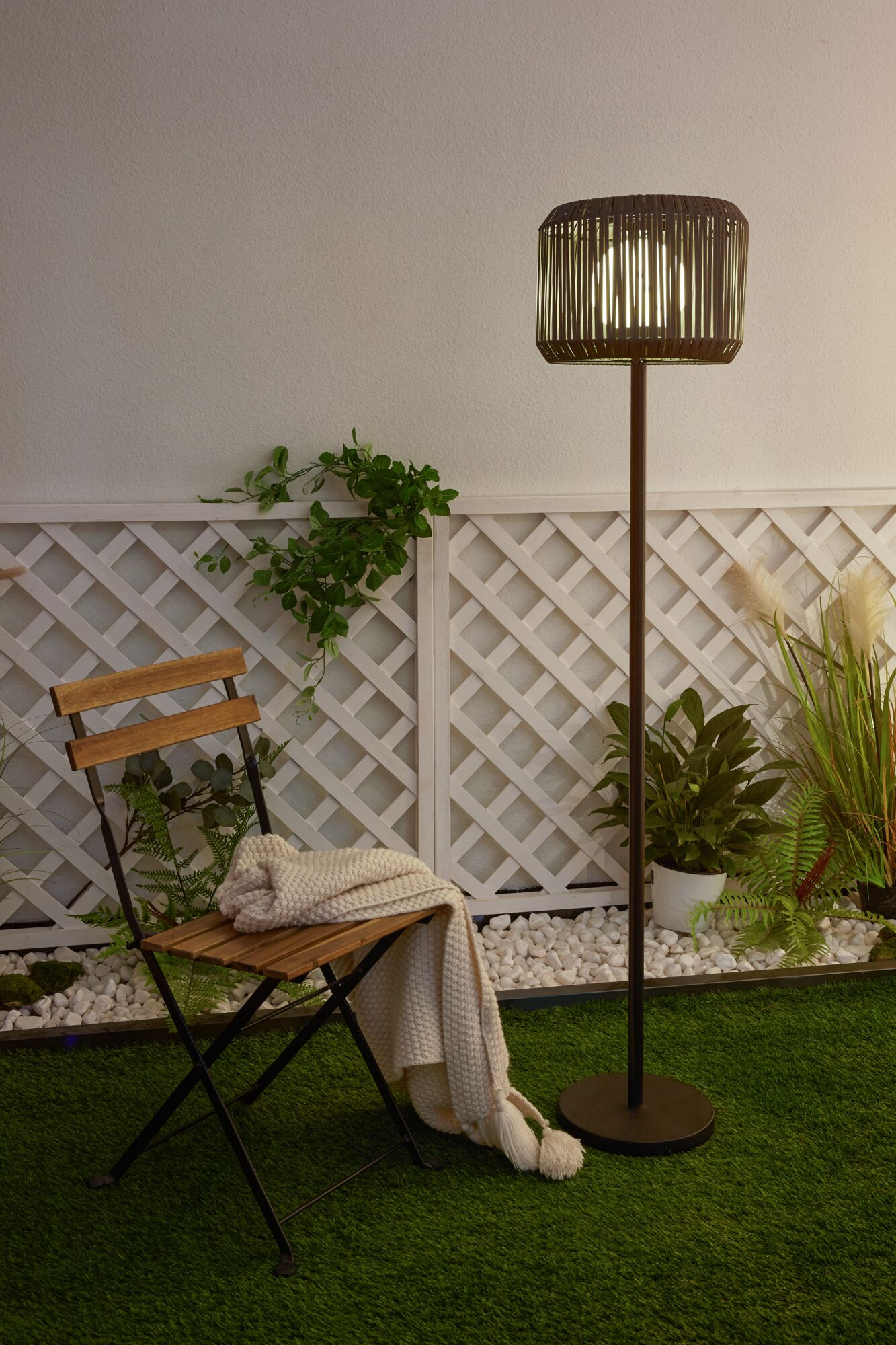 Eine Stehlampe steht auf Rasen neben einem Stuhl mit Decke vor einem Gartenzaun mit Pflanzen.
