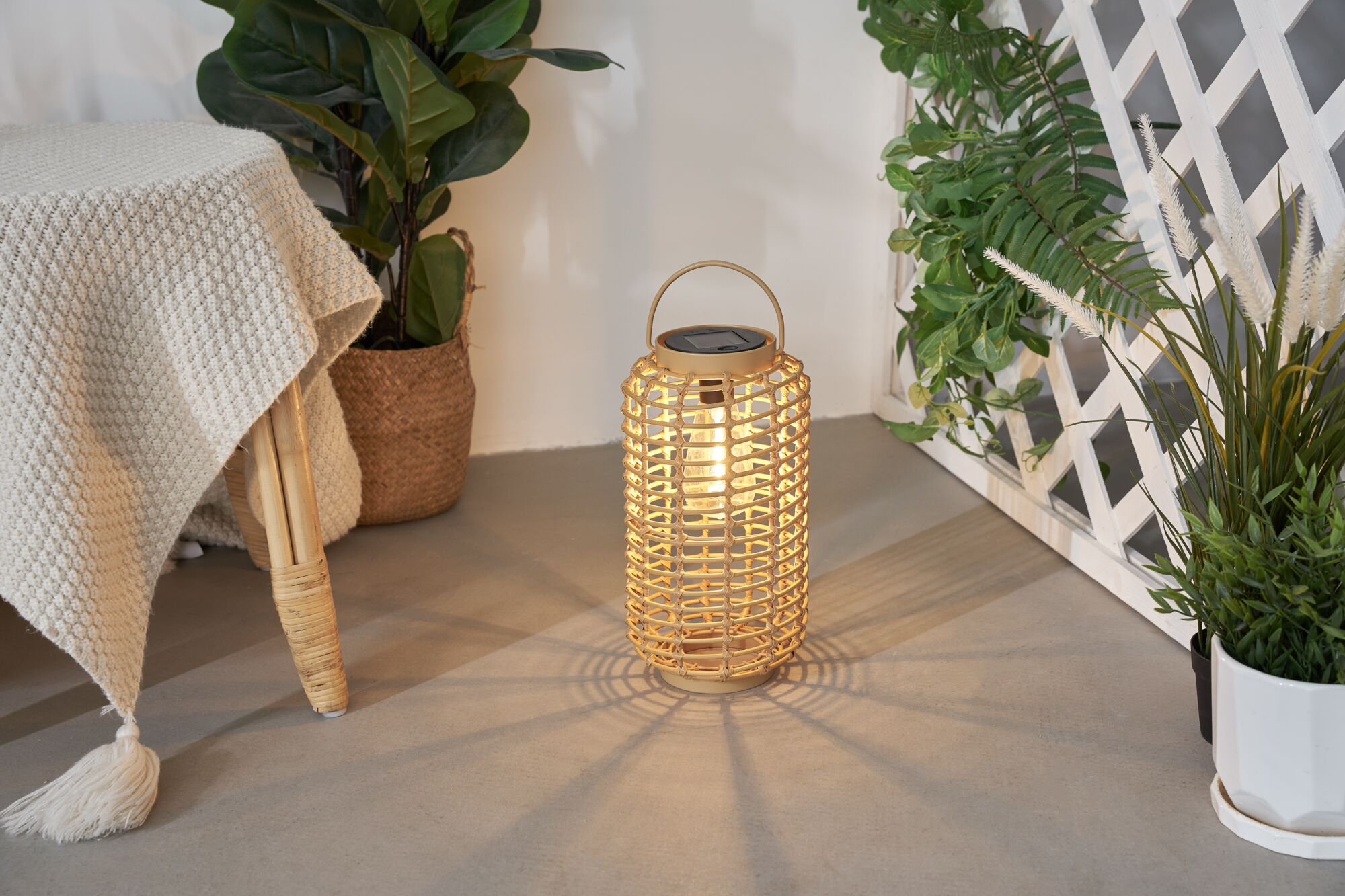 Eine Rattanlaterne leuchtet auf einem grauen Boden, umgeben von Pflanzen und einem gedeckten Tisch.