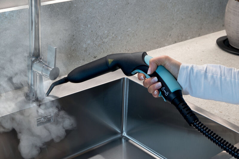Hand hält Dampfreiniger, der Dampf auf eine Küchenspüle und einen Wasserhahn sprüht.