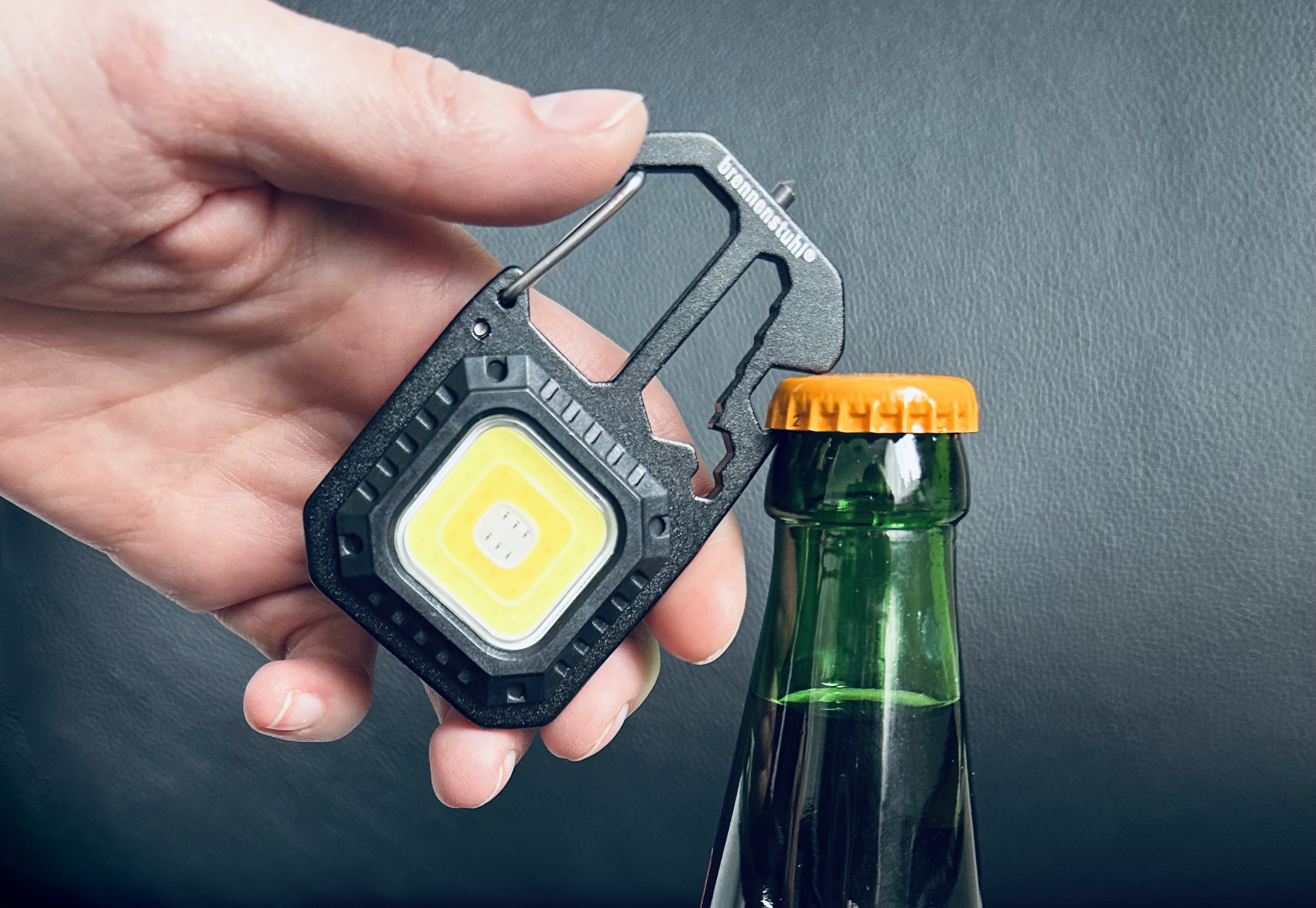 Eine Hand hält eine schwarze Mehrzweck-Taschenlampe mit Karabiner, die kurz davor ist, eine Bierflasche zu öffnen.