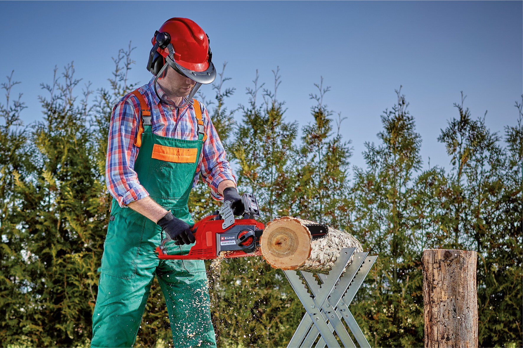 Ein Arbeiter in Schutzkleidung schneidet draußen einen Holzstamm mit einer Kettensäge.