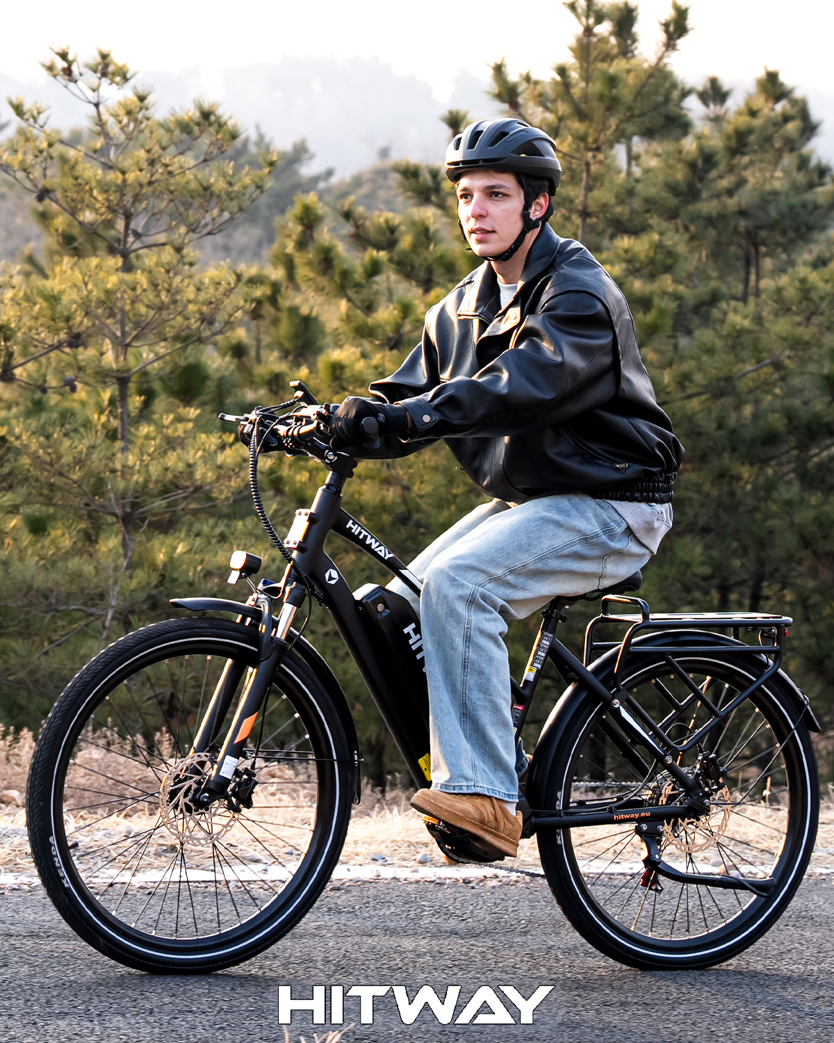 Ein junger Mann mit Helm und Lederjacke fährt auf einem schwarzen E-Bike auf einer Straße vor einer bewaldeten Hügellandschaft.