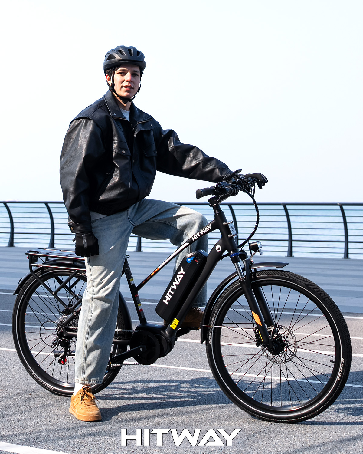 Ein junger Mann mit Helm und Lederjacke steht neben einem Hitway E-Bike auf einer Promenade.