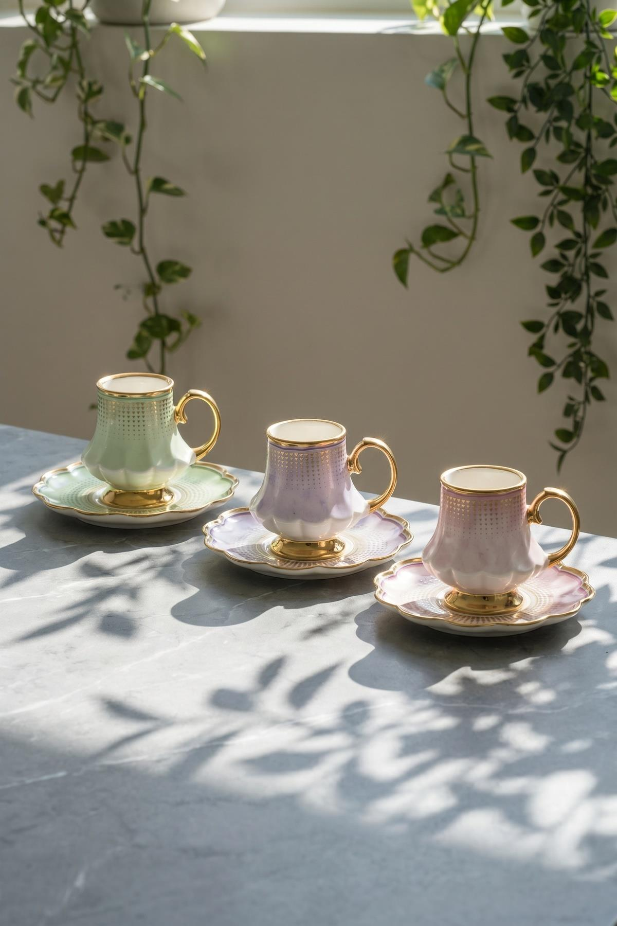 Altın kenarlı ve tabaklı üç pastel fincan mermer masada duruyor, arkada sarkan bitkiler var.