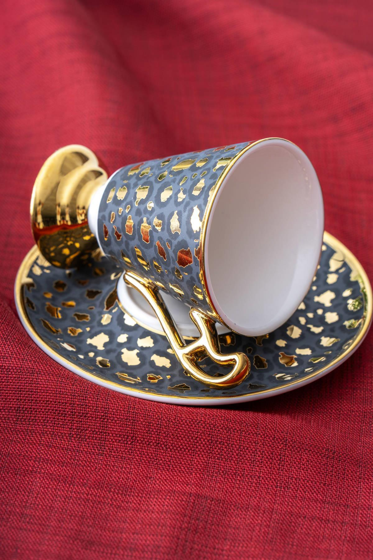 Altın leke desenli ve altın kulplu gri bir kahve fincanı, kırmızı kumaş arka plan üzerinde uyumlu bir tabakta duruyor.