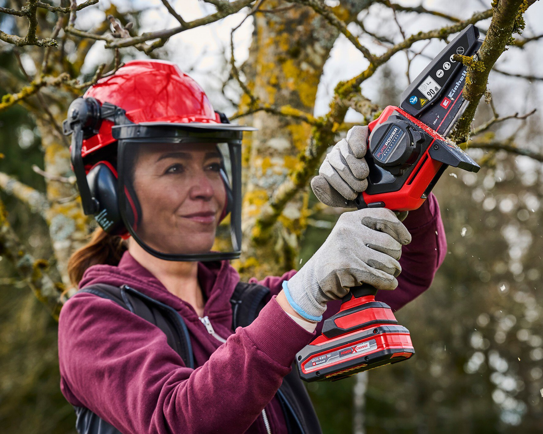Frau mit Schutzhelm und Handschuhen schneidet einen Ast mit einer roten Akku-Kettensäge.