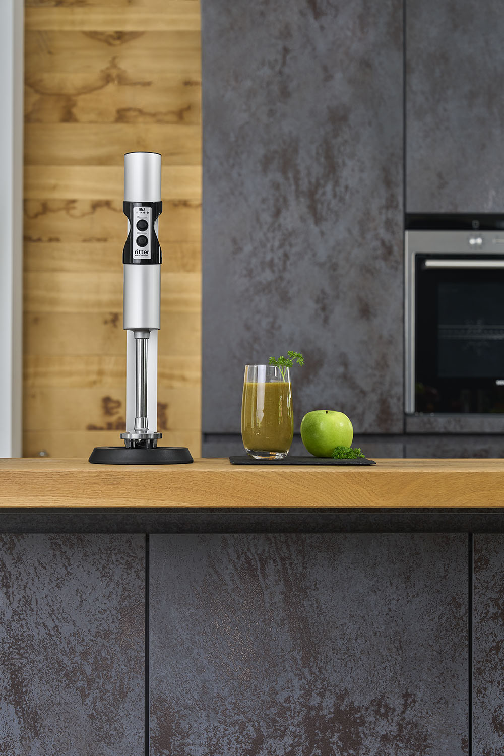 Mutfaktaki ahşap tezgah üzerinde yeşil smoothie ve bir elmanın yanında gümüş renkli bir el blenderı duruyor.