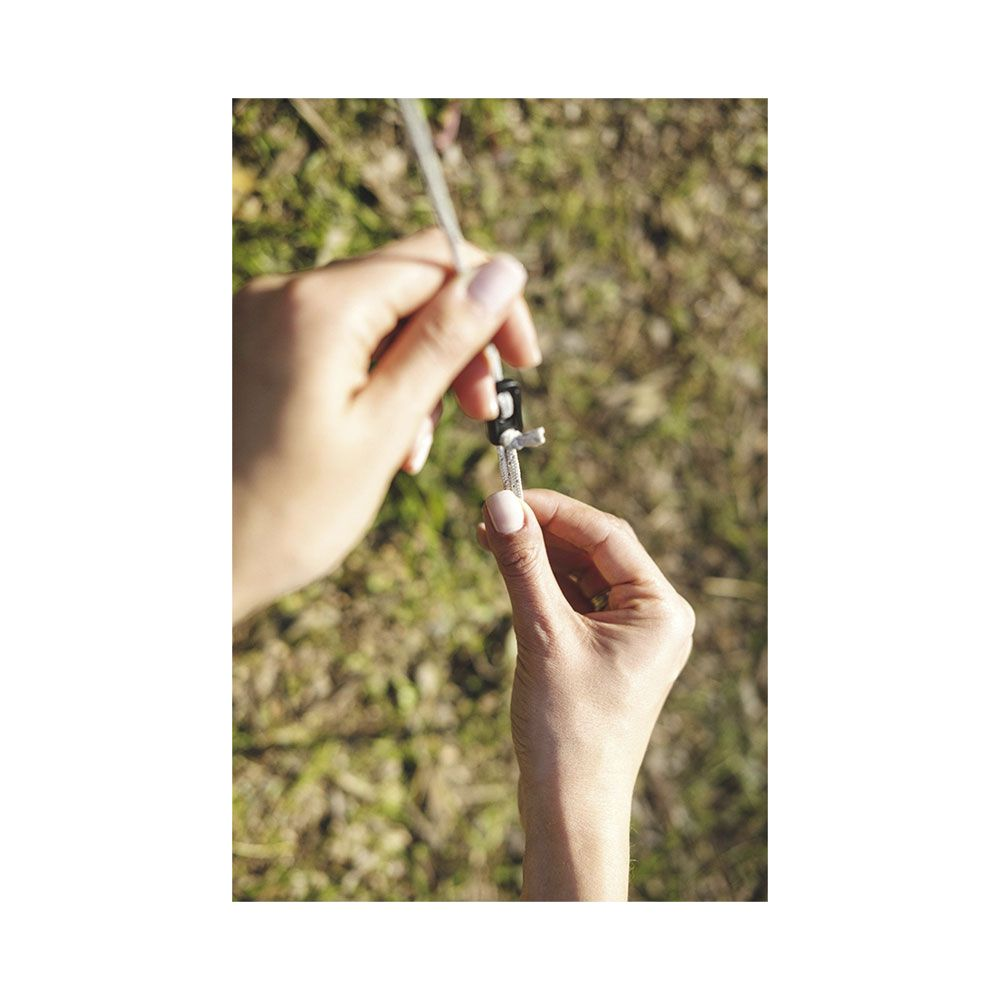 Des mains ajustent un tendeur de hauban sur une corde de tente sur de l'herbe verte.
