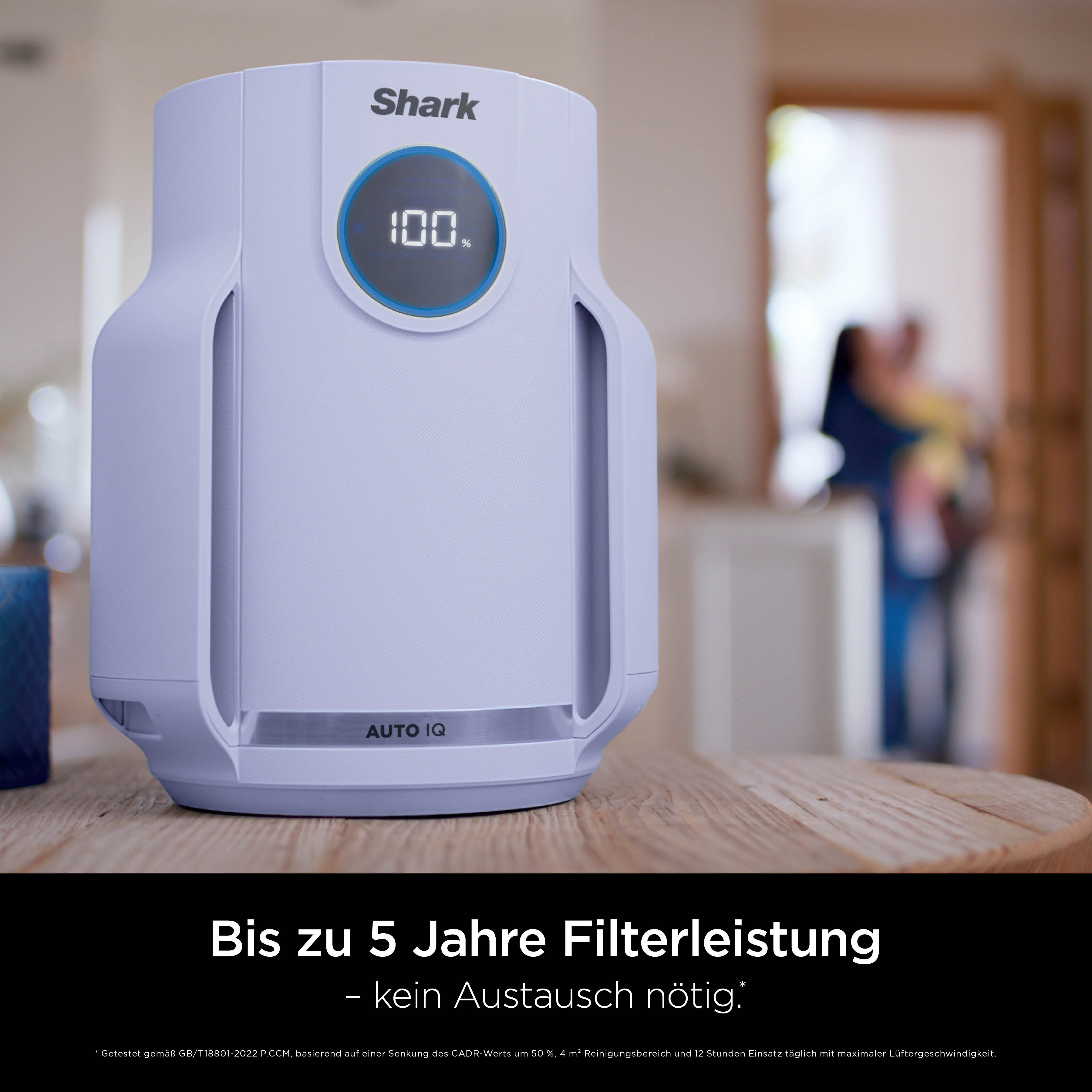 Ein weißer Shark Luftreiniger steht auf einem Holztisch, im Hintergrund ist verschwommen ein Wohnraum zu sehen.