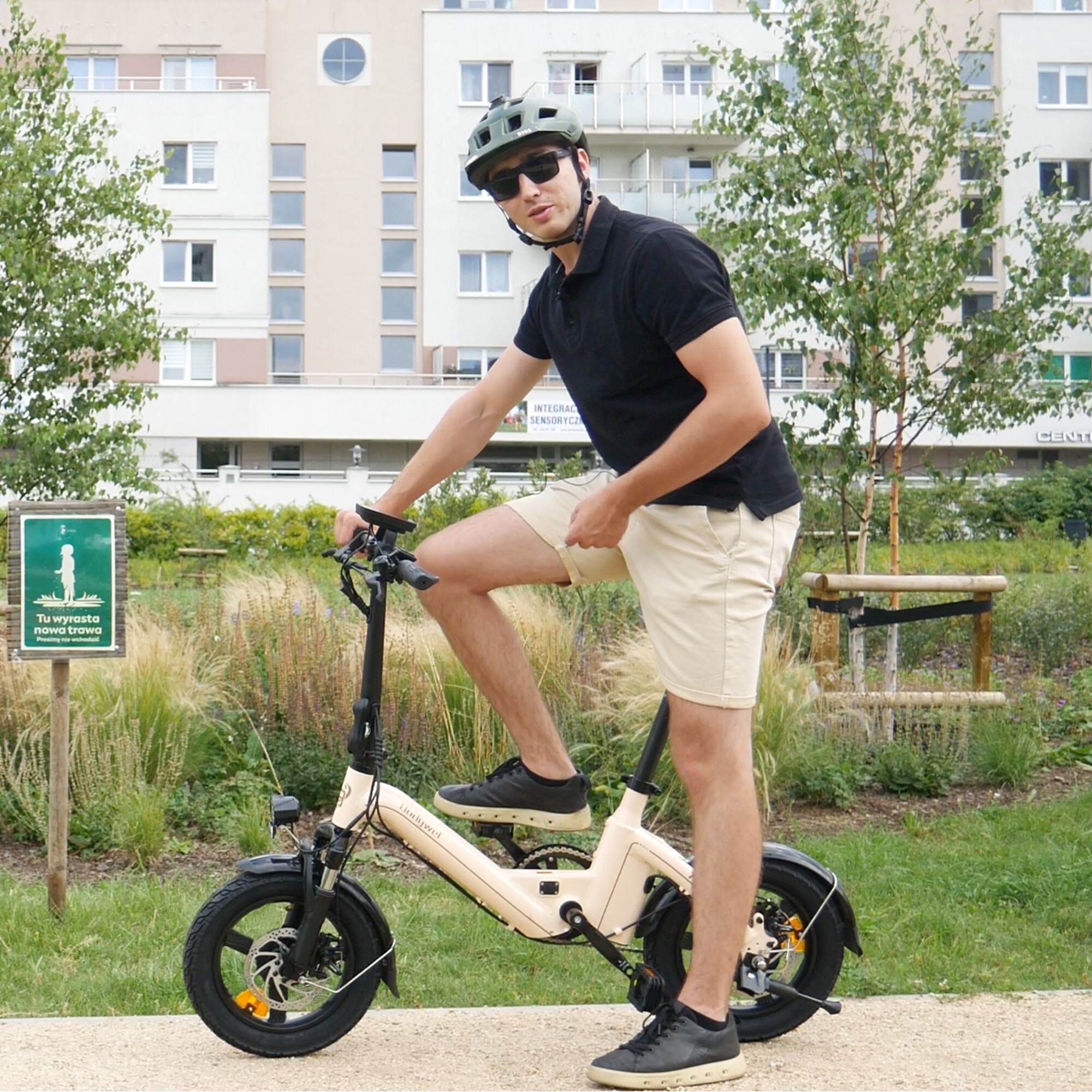 Mężczyzna w kasku i okularach przeciwsłonecznych pozuje na beżowym rowerze elektrycznym.