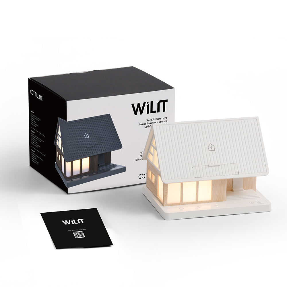 Eine weiße hausförmige Schreibtischlampe mit beleuchteten Fenstern, daneben eine Produktverpackung und ein Manual.