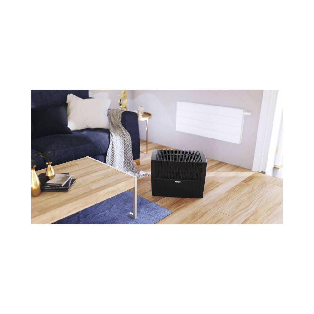 Ein schwarzer Luftbefeuchter steht auf einem Holzfußboden in einem gemütlichen Wohnzimmer mit Sofa und Couchtisch.
