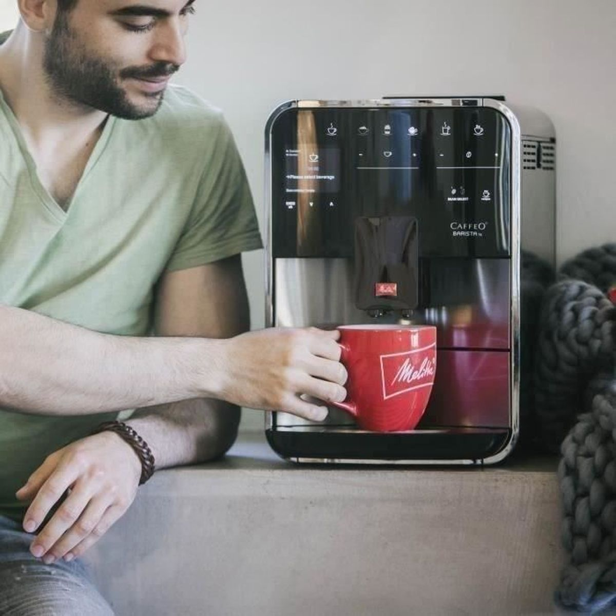 Un uomo con una maglietta verde tiene una tazza rossa davanti a una moderna macchina da caffè.