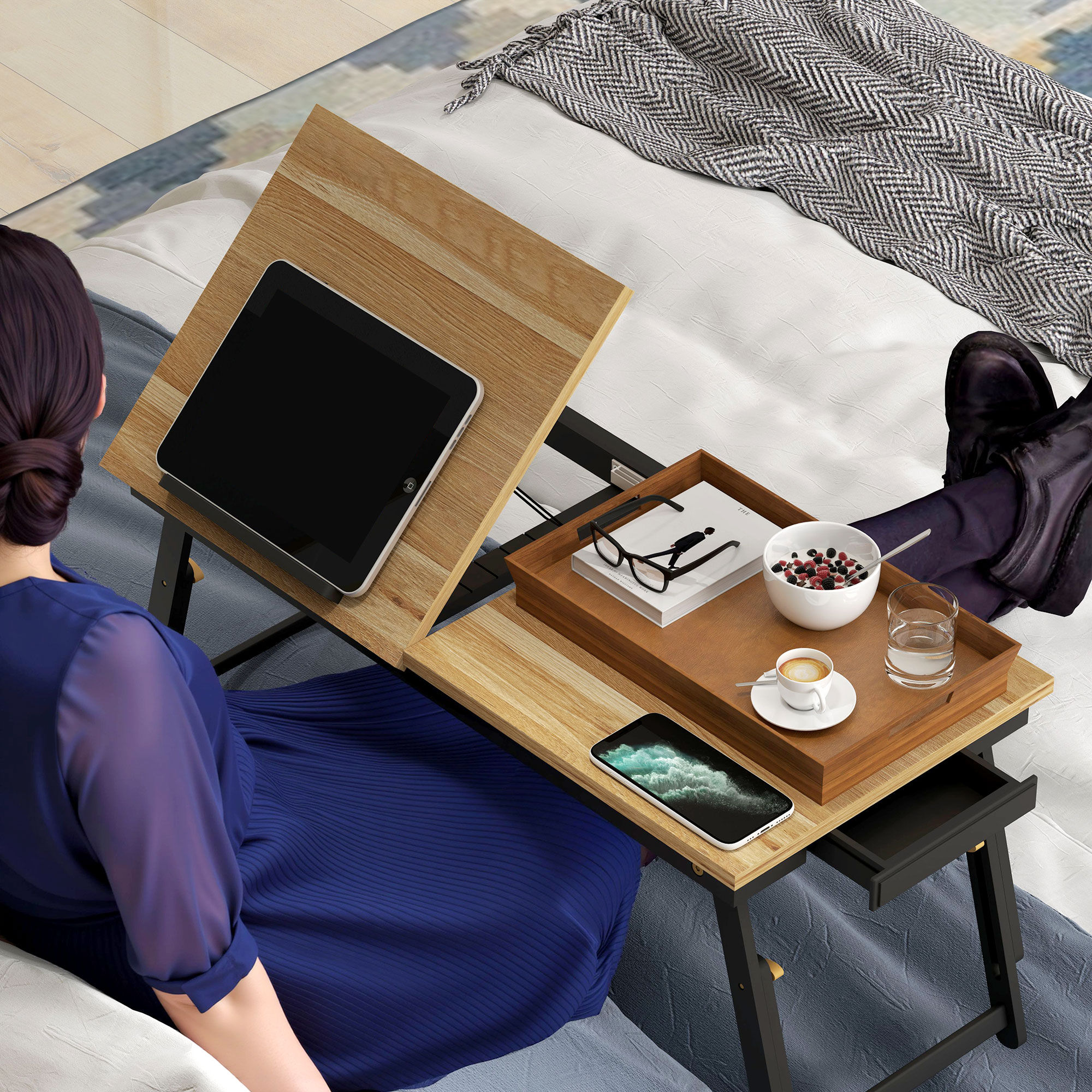 Una mujer con vestido azul está sentada en la cama usando una bandeja de cama de madera con una tableta, libro, bayas y café.