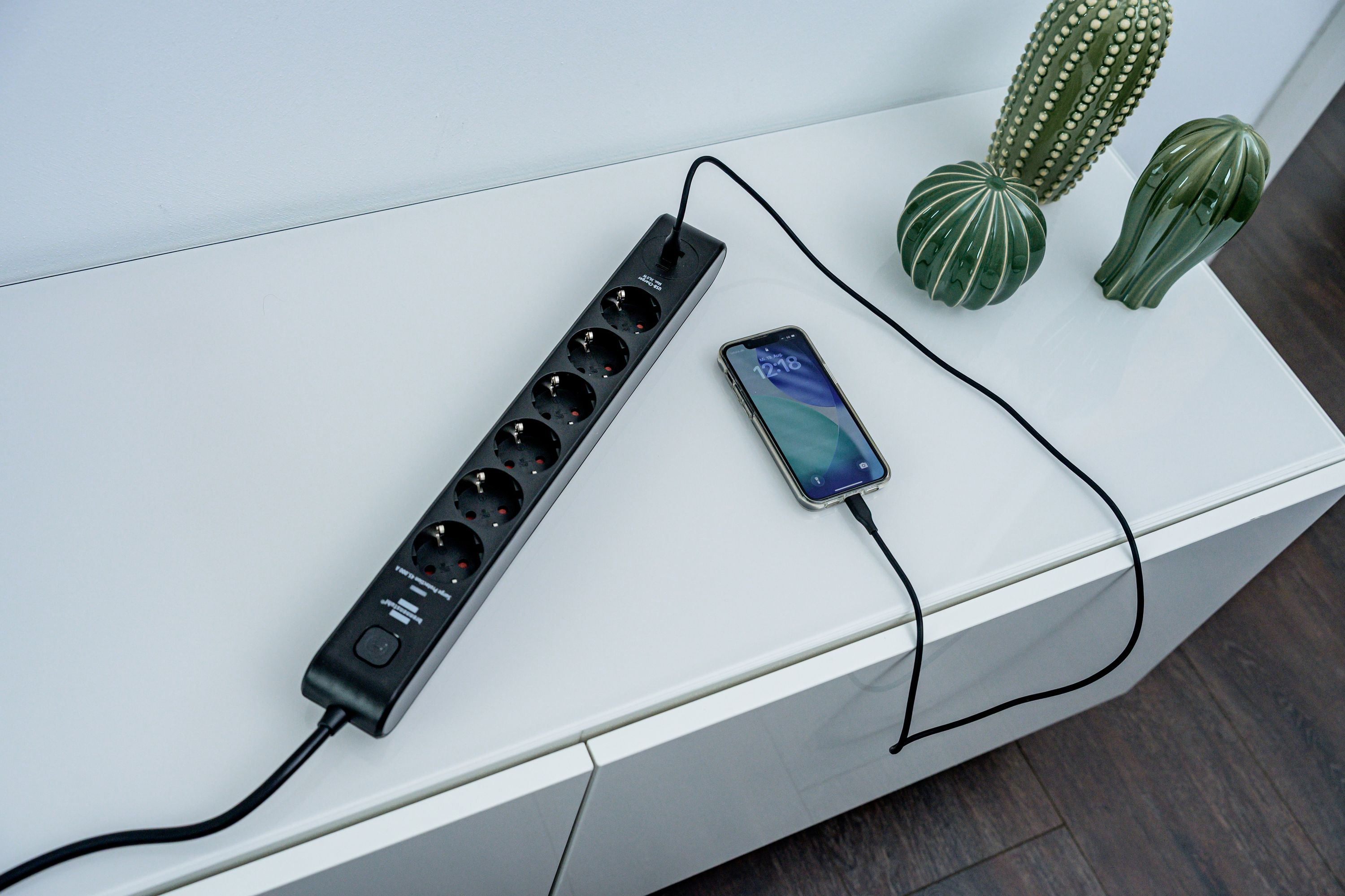 Eine schwarze Steckdosenleiste auf einer weißen Kommode, an der ein Smartphone angeschlossen ist. Drei Deko-Kaktusse stehen daneben.