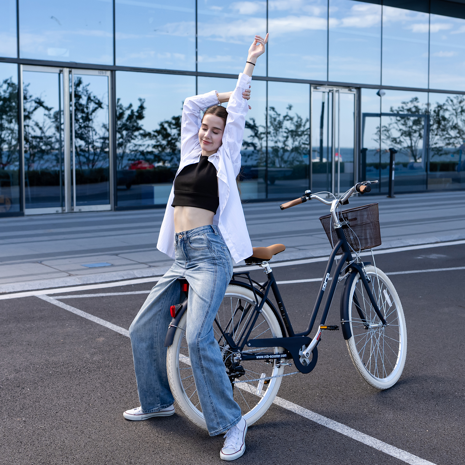 Eine lächelnde junge Frau in schwarzem Crop-Top und Jeans streckt die Arme neben einem Fahrrad im Freien.