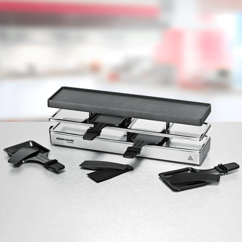 Ein silberner Raclette-Grill mit schwarzer Grillplatte und Pfännchen steht auf einer Metalloberfläche.