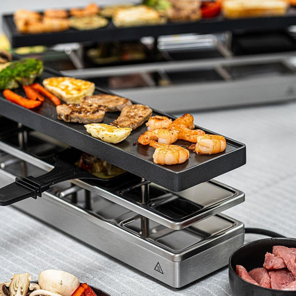 Ein Raclette-Grill gart Fleisch, Shrimps und Gemüse auf oberen Platten, mit rohen Zutaten daneben.